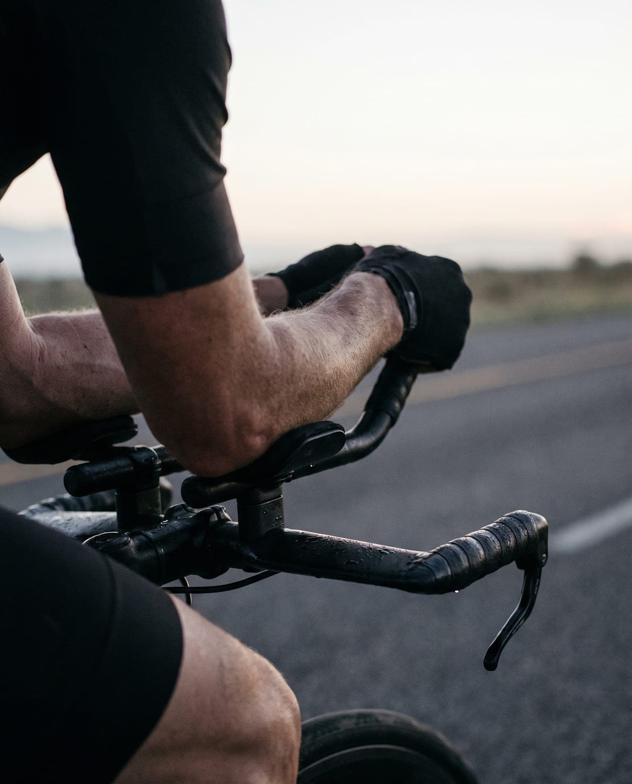 A cyclist's forearms and hands rest on the aero bars of a time-trial bike at dawn, the road ahead blurred in pale light.
